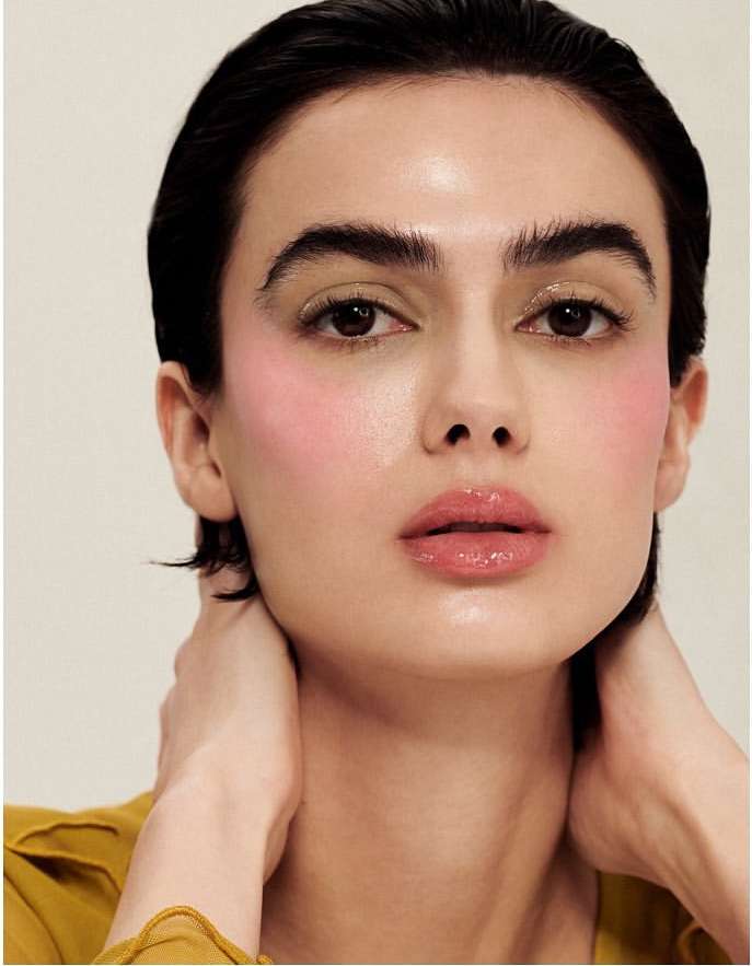 Close-up portrait of a woman with short dark hair and pink blush, wearing a yellow shirt, against a neutral background.