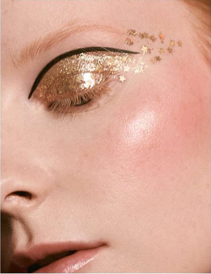 Close-up of an adult female eye with gold glitter eyeshadow, black eyeliner, and gold star-shaped confetti on fair skin.