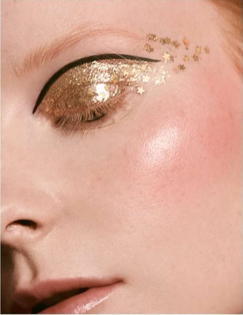 Close-up of an adult female eye with gold glitter eyeshadow, black eyeliner, and gold star-shaped confetti on fair skin.
