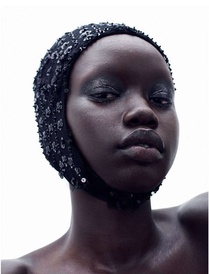 Close-up portrait of a Black woman with dark eyeshadow wearing a beaded black head covering, set against a bright, neutral background.