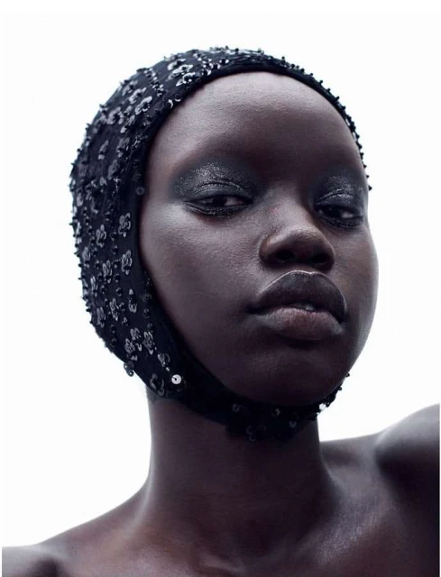 Close-up portrait of a Black woman with dark eyeshadow wearing a beaded black head covering, set against a bright, neutral background.