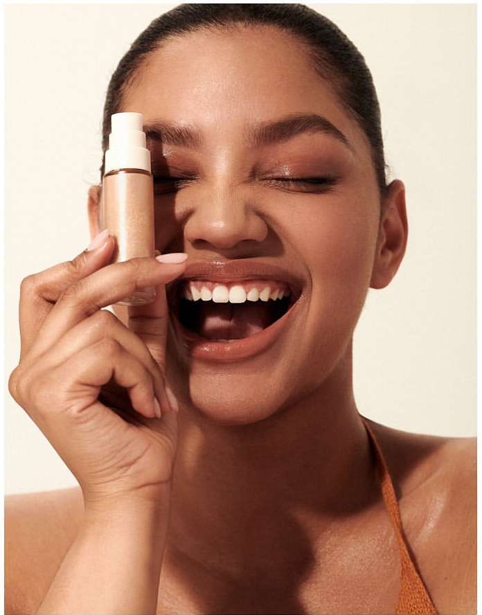 Radiant woman with closed eyes smiling while holding a bottle of bronzer makeup in a beauty shot with soft neutral tones and natural light.