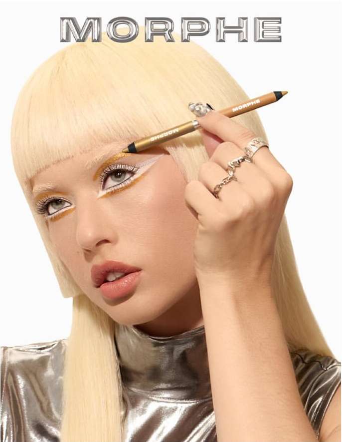 Blonde woman applies gold eyeliner in a beauty shot with a white backdrop, emphasizing makeup and a futuristic silver top under studio lighting.