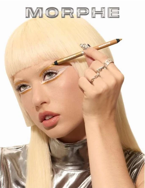 Blonde woman applies gold eyeliner in a beauty shot with a white backdrop, emphasizing makeup and a futuristic silver top under studio lighting.