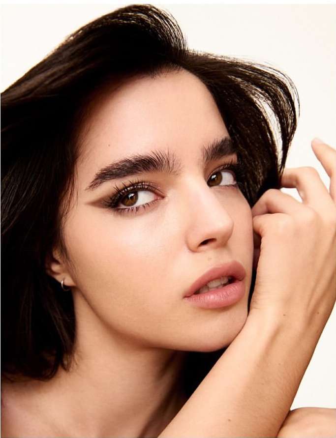 Close-up portrait of a woman with dark hair and brown eyes, showcasing winged eyeliner and subtle nude lipstick against a soft white background.