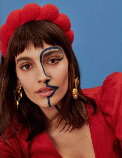 Close-up of a woman with brown hair, wearing a red headband and gold earrings, with a blue abstract design painted on half her face.