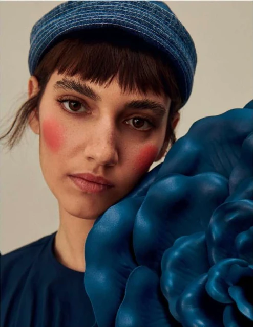 Close-up of a woman with brown hair wearing a denim hat and dark-blue top with a large teal floral prop alongside her face.
