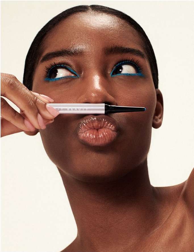 A young Black woman with bright blue eyeliner poses with an eyeliner pencil over her lips in a beauty close-up, against a white backdrop.