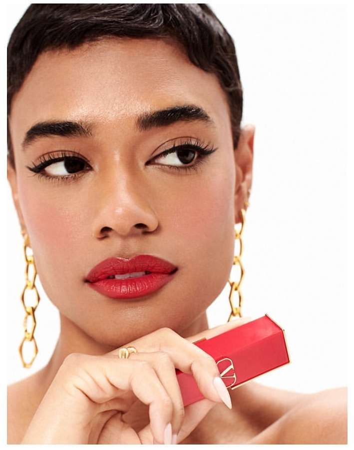 Close-up of a woman with short dark hair and gold chain earrings, red lipstick, and holding a red lipstick tube, white background.