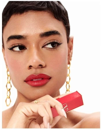 Close-up of a woman with short dark hair and gold chain earrings, red lipstick, and holding a red lipstick tube, white background.