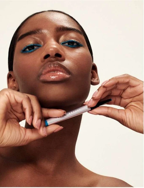 Close-up of a dark-skinned woman applying bright blue eyeliner with a makeup pencil in a beauty or fashion editorial.