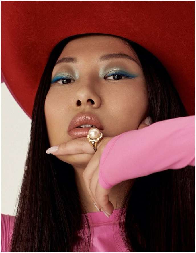 Portrait of an Asian woman with dramatic makeup, bright pink top, and a large red hat, wearing a large pearl ring.