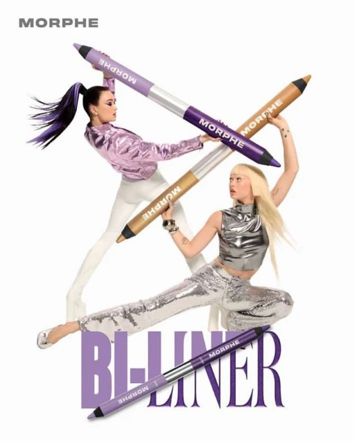 Two women pose creatively with oversized Morphe B-liner makeup pencils in a studio shot against a white background. Beauty product promotion.