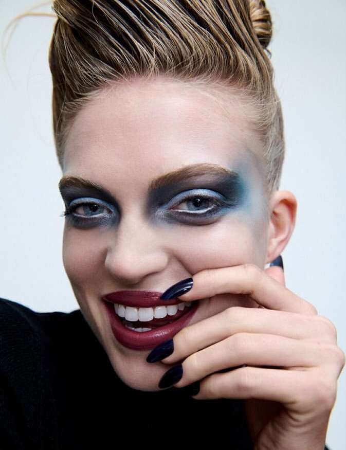 Close-up portrait of woman with blue eyeshadow, burgundy lipstick, and dark long nails near her face against a white background.