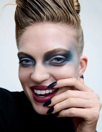 Close-up portrait of woman with blue eyeshadow, burgundy lipstick, and dark long nails near her face against a white background.