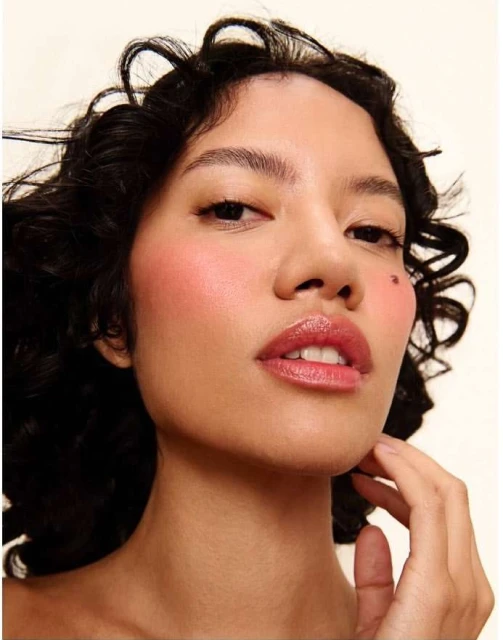 Close-up portrait of a woman with curly dark hair and light skin, featuring pink blush and shiny lips against a neutral background.