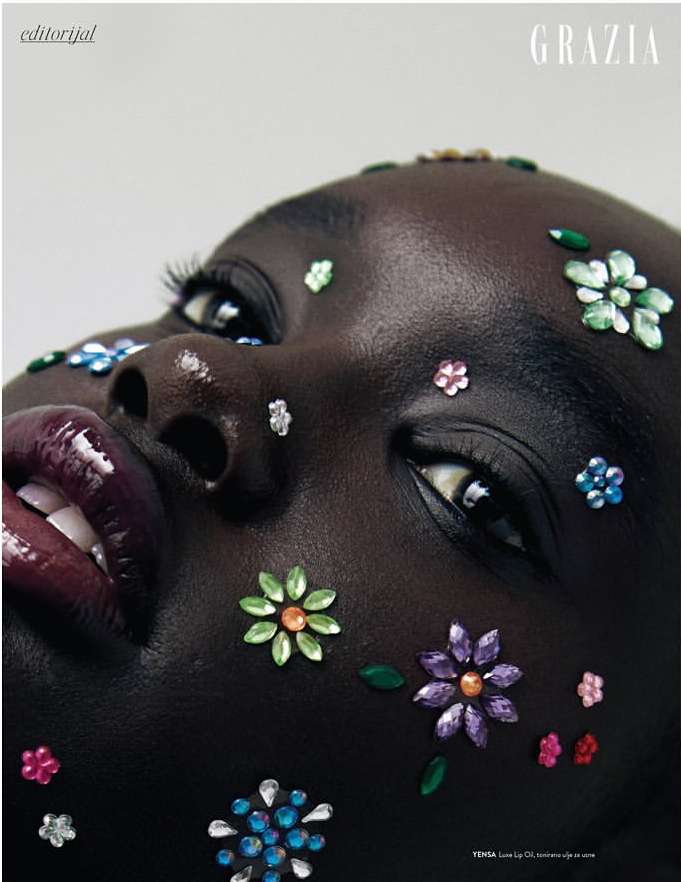 Close-up of an adult female model with dark skin, flower-shaped gem makeup, glossy lipstick, and white teeth against a neutral background.