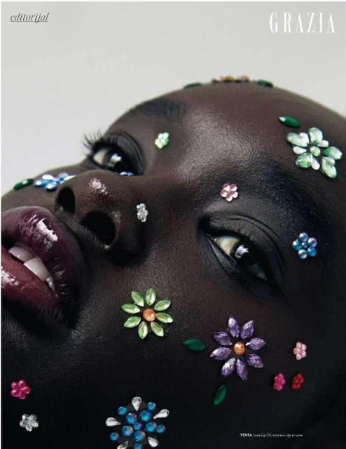 Close-up of an adult female model with dark skin, flower-shaped gem makeup, glossy lipstick, and white teeth against a neutral background.