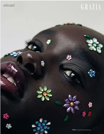 Close-up of an adult female model with dark skin, flower-shaped gem makeup, glossy lipstick, and white teeth against a neutral background.