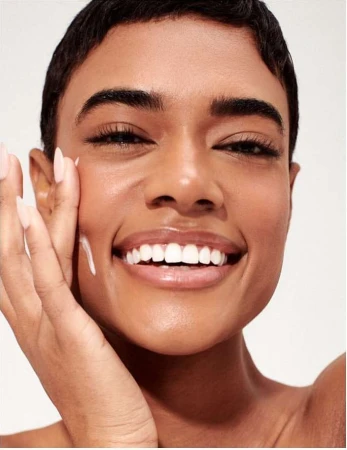 Close-up of a smiling African American woman applying cream to her face with a light background, showcasing skincare routine.