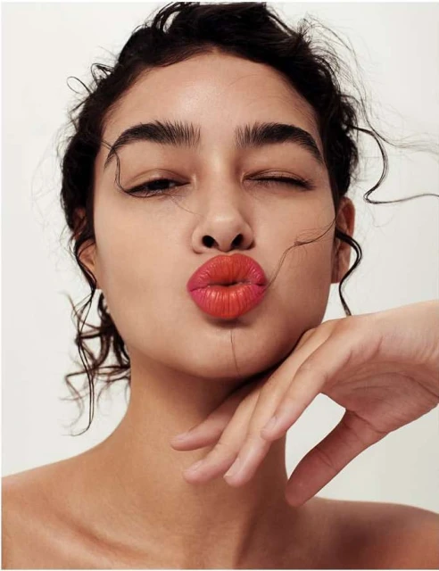 Close-up studio portrait of a young woman with curly hair winking and puckering her lips, showcasing bold red lipstick against a white backdrop.