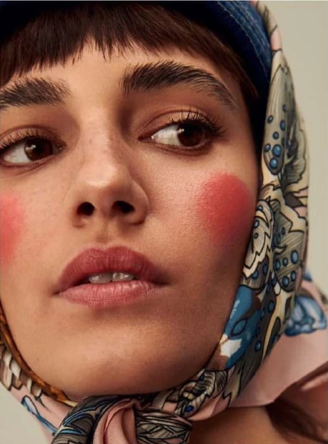 Close-up shot of a young adult female model with fair skin, wearing a patterned headscarf and noticeable rouge makeup on her cheeks.