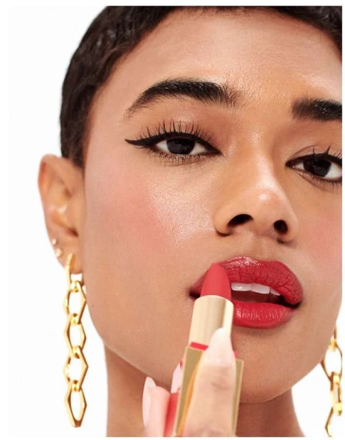 Close-up of a woman with dark hair applying red lipstick; gold earrings are visible against a white background, creating a beauty aesthetic.