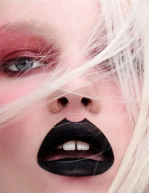 Close-up of a woman's face with stark makeup: bold black lipstick and pink eyeshadow, with light blonde hair partially obscuring her face.