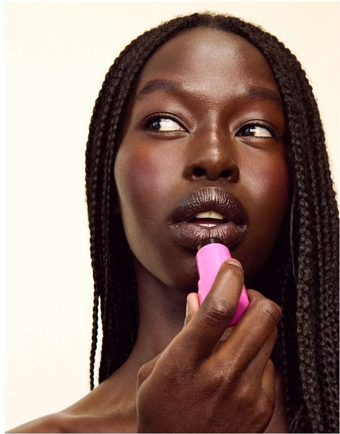 Portrait of a dark-skinned woman applying pink lipstick, showcasing braided hair, and a makeup look against a neutral backdrop.