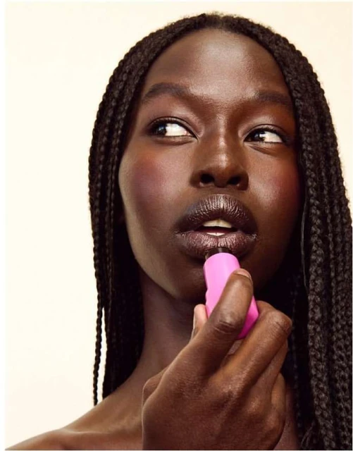 Portrait of a dark-skinned woman applying pink lipstick, showcasing braided hair, and a makeup look against a neutral backdrop.