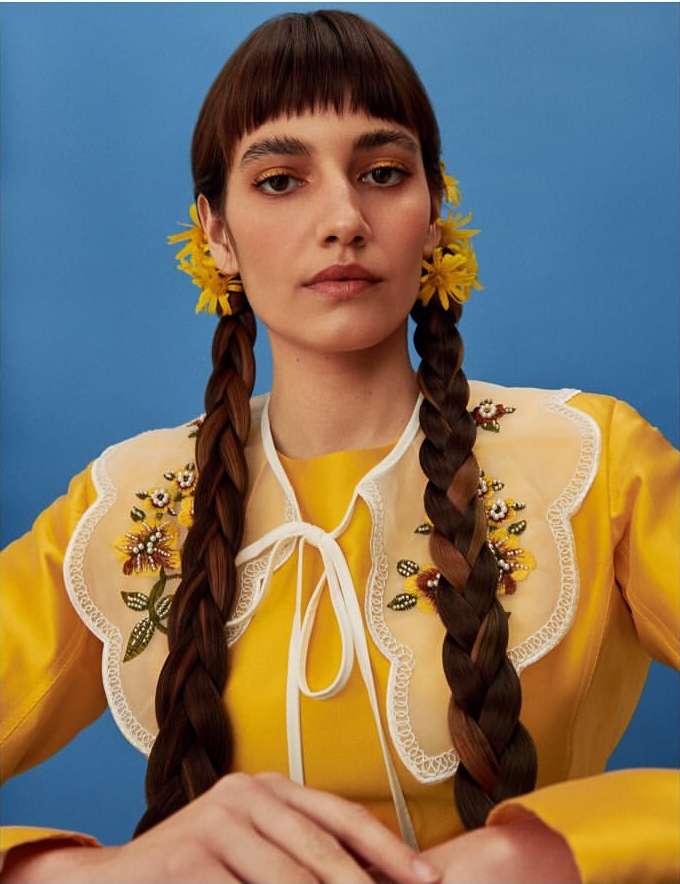 Portrait of a young adult woman with yellow flowers in her braided pigtails, wearing a yellow top with an embellished collar, against blue.
