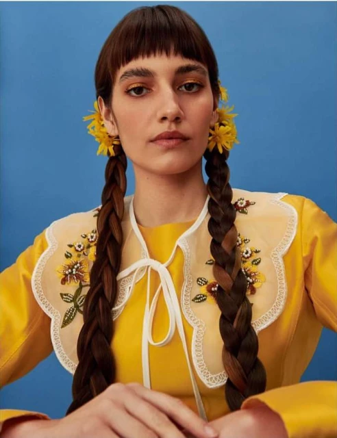Portrait of a young adult woman with yellow flowers in her braided pigtails, wearing a yellow top with an embellished collar, against blue.