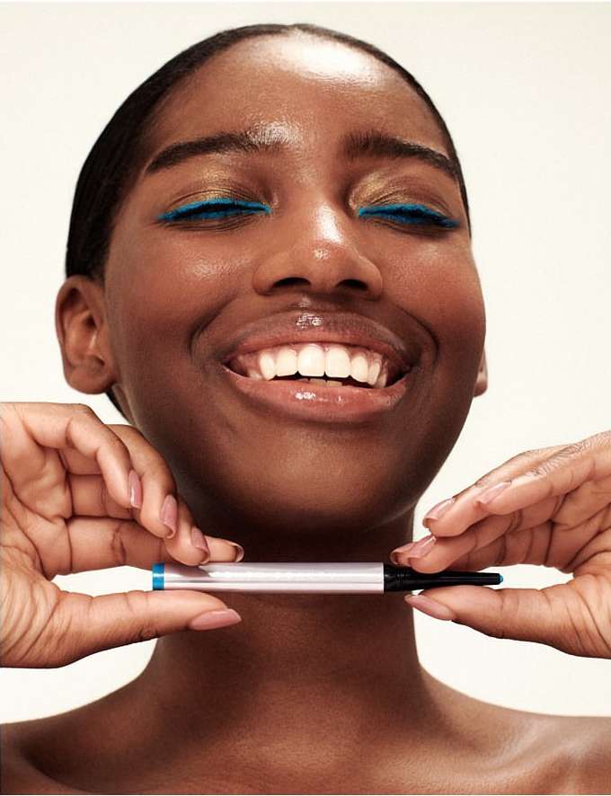 Close-up of a smiling Black woman with blue eyeliner, holding an eyeliner pencil, illuminated by bright indoor lighting on a white backdrop.