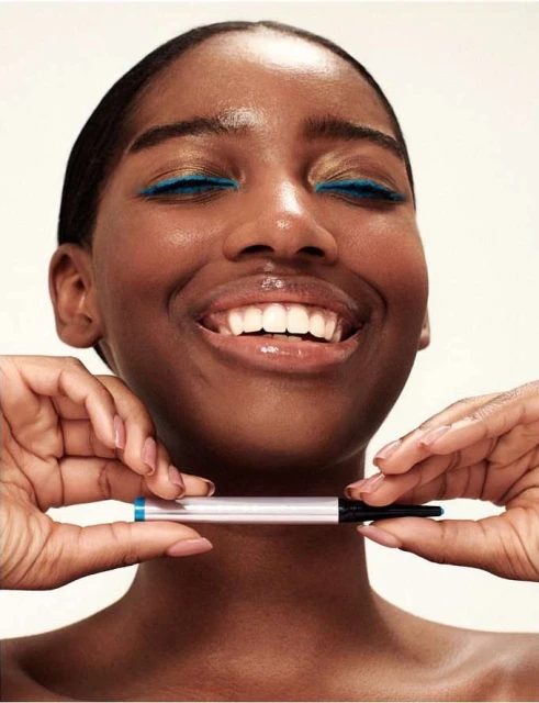 Close-up of a smiling Black woman with blue eyeliner, holding an eyeliner pencil, illuminated by bright indoor lighting on a white backdrop.
