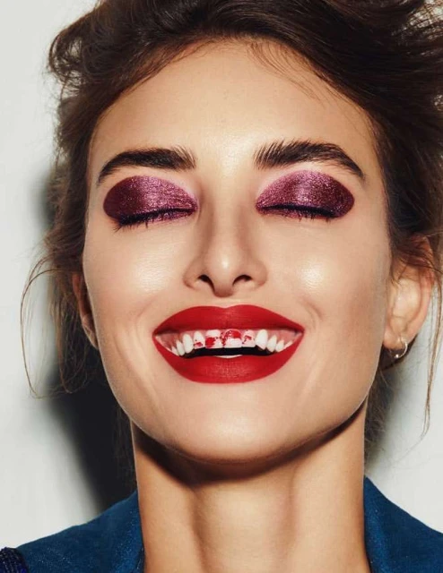 Close-up of a smiling woman with dramatic red lipstick and dark purple eye shadow against a white background, beauty and makeup concept.