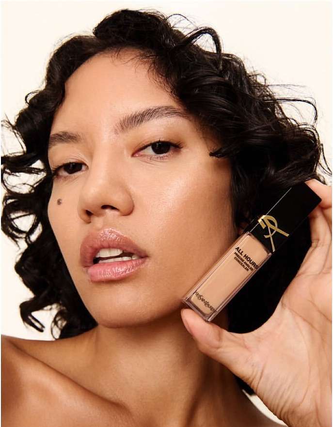 Woman holding YSL All Hours Precise Angles Concealer. Close-up of a person with dark curly hair, light skin, and glossy lips.