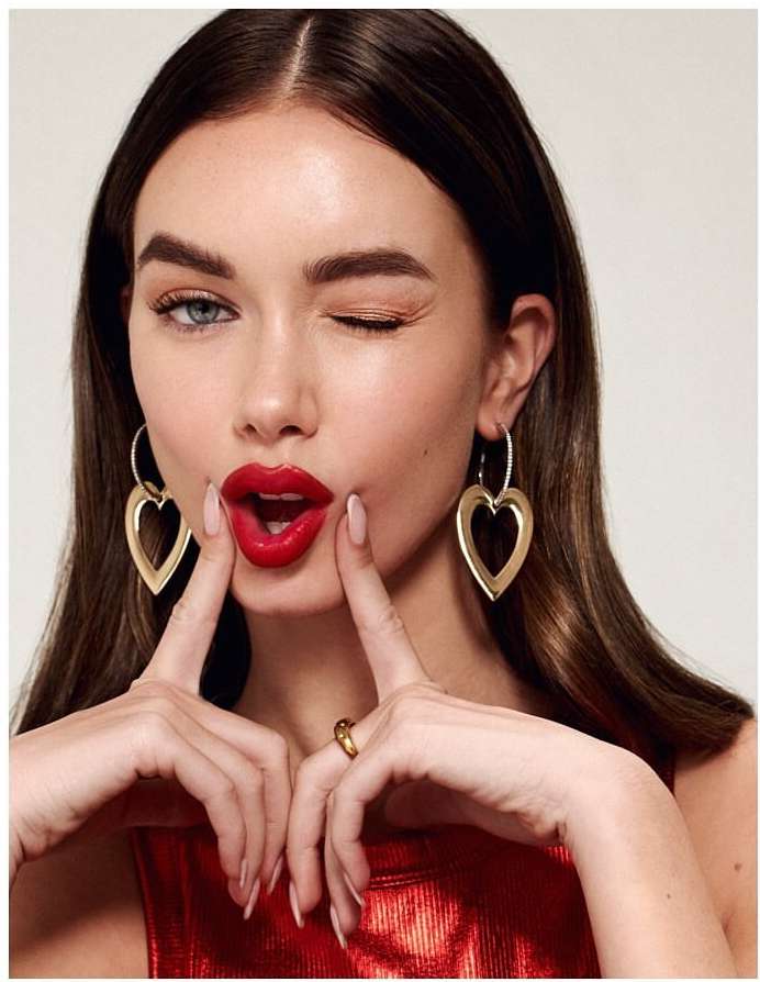 Portrait of a woman with bright red lipstick winking and wearing heart-shaped earrings and a red metallic shirt in a bright studio shot.