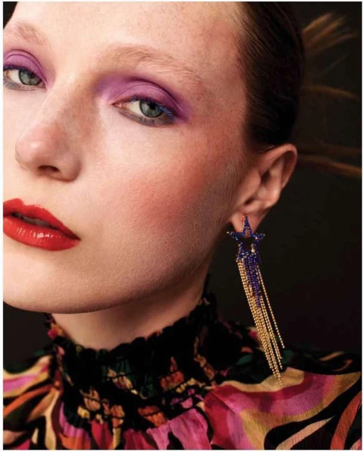 Close-up of a red-lipsticked woman with lavender eyeshadow, wearing star-shaped earrings with gold and blue tassels, fashion shot.