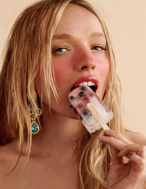 Close-up of a fair-skinned woman with long blonde hair eating a fruit popsicle against a peach-colored background, wearing statement earrings.