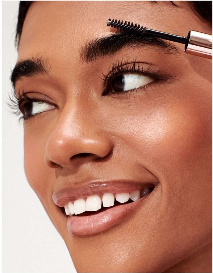 Close-up of a smiling woman with dark hair applying eyebrow gel with a wand, showcasing beauty and personal care product usage.