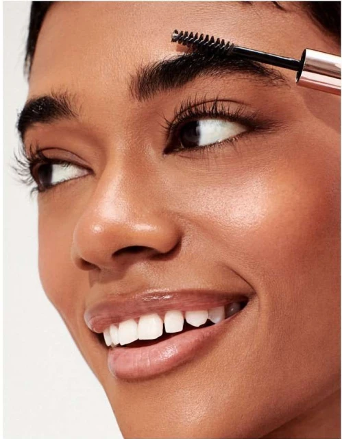 Close-up of a smiling woman with dark hair applying eyebrow gel with a wand, showcasing beauty and personal care product usage.