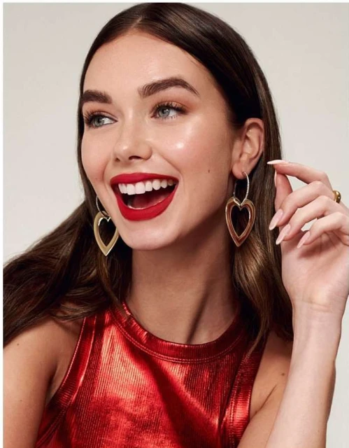 Smiling woman with red lipstick and red top poses with heart earrings against a neutral backdrop in a beauty shot.