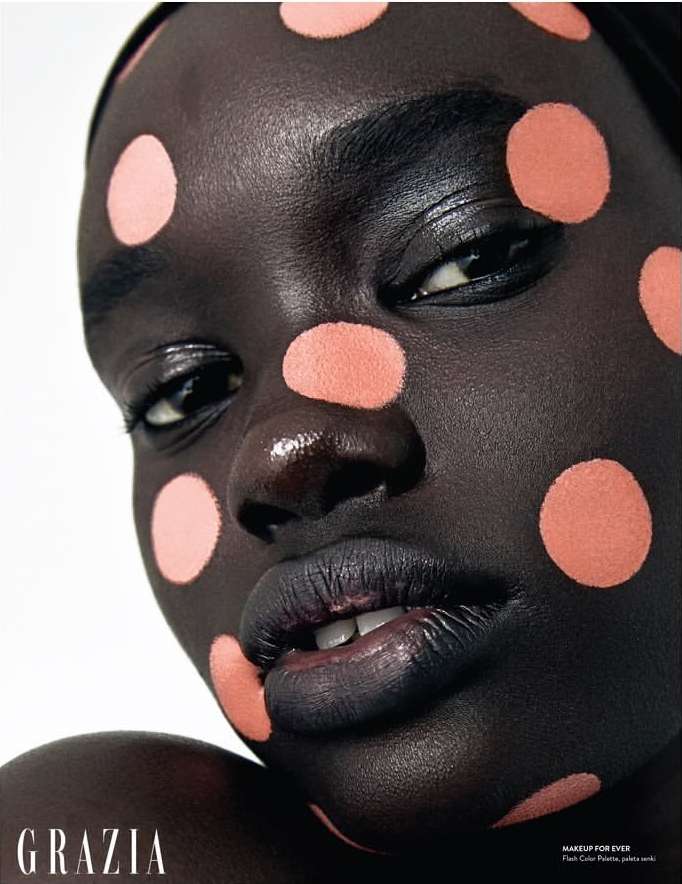 Close-up beauty shot of a woman with dark skin wearing peach-colored polka dots on her face and dark lipstick, with 'Grazia' logo.