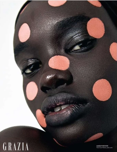 Close-up beauty shot of a woman with dark skin wearing peach-colored polka dots on her face and dark lipstick, with 'Grazia' logo.