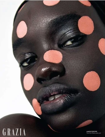 Close-up beauty shot of a woman with dark skin wearing peach-colored polka dots on her face and dark lipstick, with 'Grazia' logo.