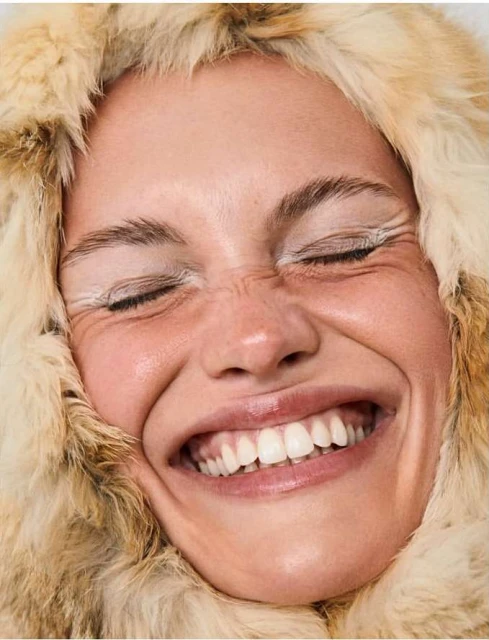 Close-up studio shot of a smiling woman with closed eyes and white eyeliner, wearing a fur hood; beauty, fashion, portrait photography.