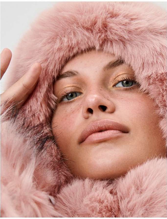 Close-up portrait of a fair-skinned woman with light eyes and freckles wearing a pink faux fur hooded coat against a white backdrop.