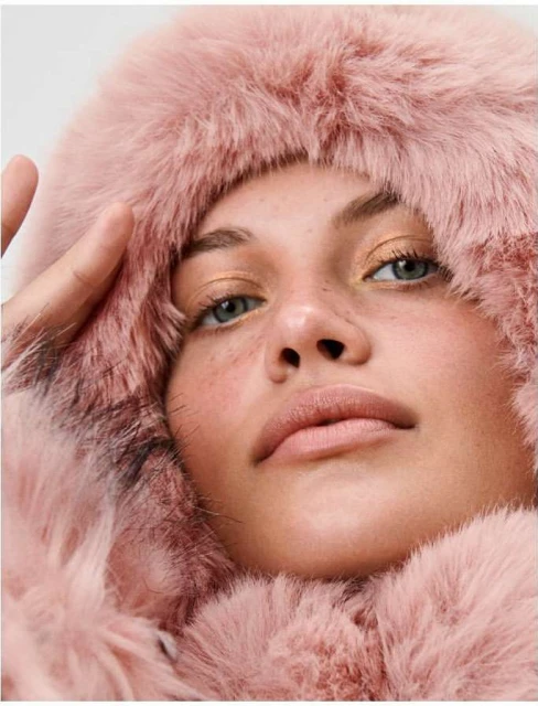 Close-up portrait of a fair-skinned woman with light eyes and freckles wearing a pink faux fur hooded coat against a white backdrop.