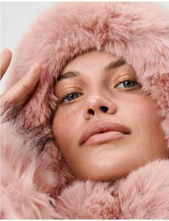Close-up portrait of a fair-skinned woman with light eyes and freckles wearing a pink faux fur hooded coat against a white backdrop.