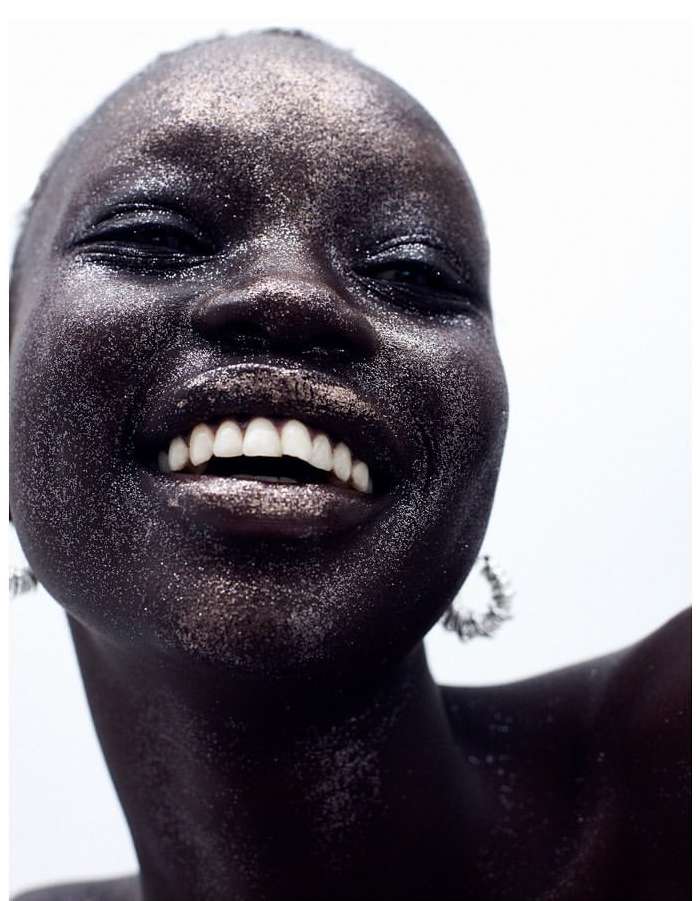 Close-up of a smiling, dark-skinned woman with glittering makeup. Her teeth are visible and she's wearing hoop earrings against a white background.